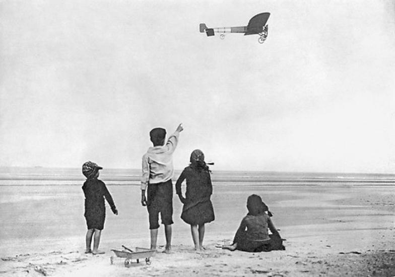 Black and white photo of four children on a beach. One points to an airplane in the distance.