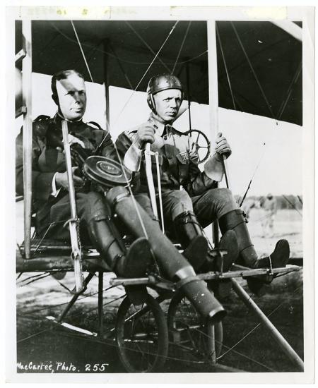 Two men in the seat of a Wright airplane. The man on the left is holding a large machine gun and the man on the right is at the controls.