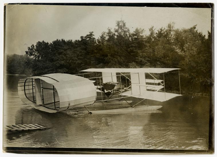 A biplane with an oval structure on the back floating on water with trees in the background.