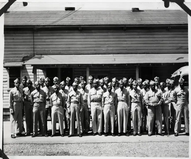 Black and white photo of approximately 30 Black men in uniform, posing in front of a wood building with an awning.