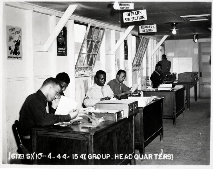 A line of four Black men work seated at a line of desks with boxes of paper and office equipment in front of them. Overhead is a sign that reads: "Officers Subsection."