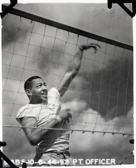 Black and white photo of a black man in a white tee shirt behind a volleyball net with his left hand in the process of spiking a ball (ball not visible)