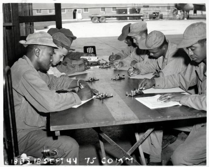 A group of Black men sit on the left and right side of a vertically place table.  They are all in uniform with various caps.  Each holds a notebook, pen, and is seated in front of a piece of a telegraph system.