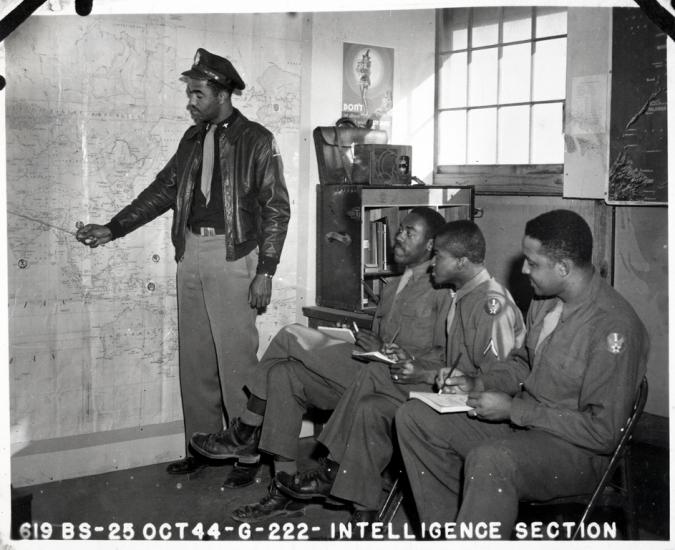 Black and white photograph. The entire back wall is a map of Europe.  A Black man in a jacket, hat, and tie points to the map with his right hand.  Three men sit in chairs to his right with notebooks and pens in hand.