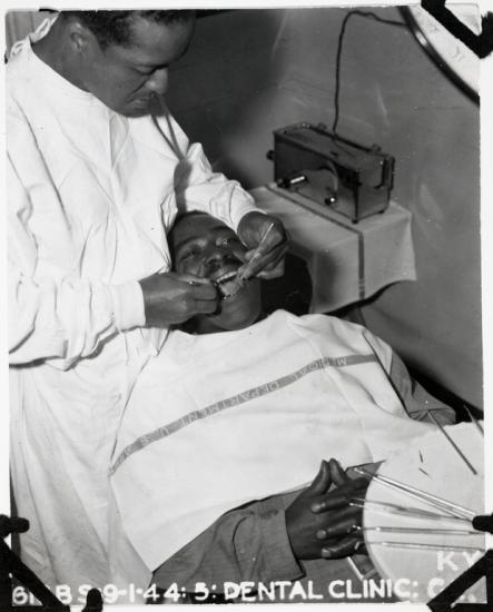 Black and White photo of a Black man standing with his dental tools in the open mouth another man, whose hands are clasped over his chest.  A tray of dental tools sits in the lower right foreground. 