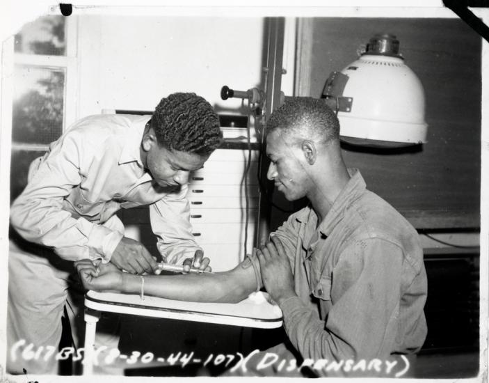 Black and white photo.  Black man on the left in a light uniform is holding a hypodermic needle to the right arm of a seated man, who is supporting his arm with his right hand.