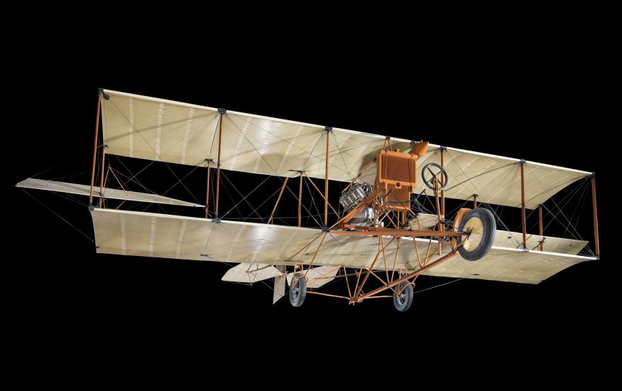 Khaki and orange colored biplane craft suspended in air against a black background.