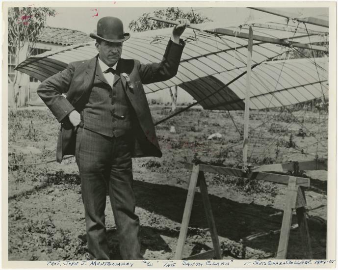 A photo of John Montgomery standing next to a glider. One hand is on the glider and the other is on his hip.