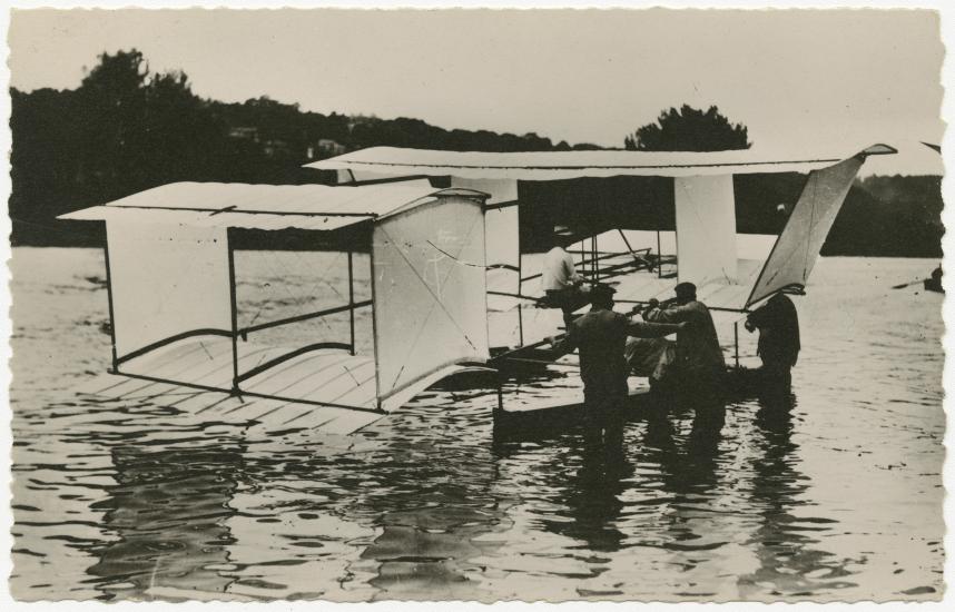 One-half right rear view of the a Blériot floatplane on the water. Men stand in the water next to it.