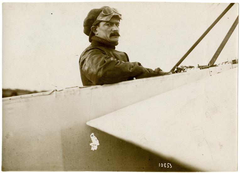 A man wearing a hat and goggles on his head, sitting in the cockpit of an airplane, looking toward the camera.