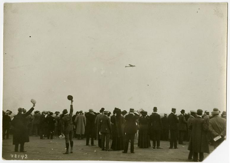 A crowd of people, some cheering, looking toward the sky where an airplane can be seen in the distance.