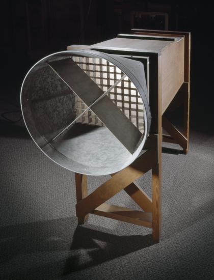 A wind tunnel with a metal circular piece attached to hollow rectangular box-shaped wind tunnel on wooden frame.