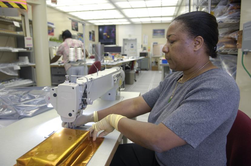 A woman sits at an industrial sewing machine, sewing a golden piece of cloth. 