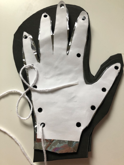 A cut out of a hand on printer paper with hole punches around the outside, stacked on top of a cut out of a hand on Mylar, stacked on top of a cut out of a glove on foam. A piece of yarn with tape around the end creating a point is threaded through the first hole.