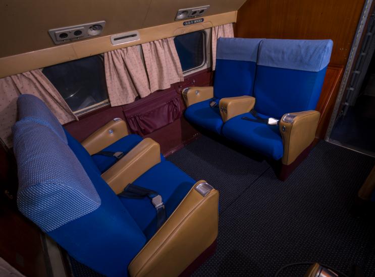 A quartet of four seats in sets of two facing each other. Behind the seats is a row of windows with curtains. The seats are blue and the surrounding walls brown. 