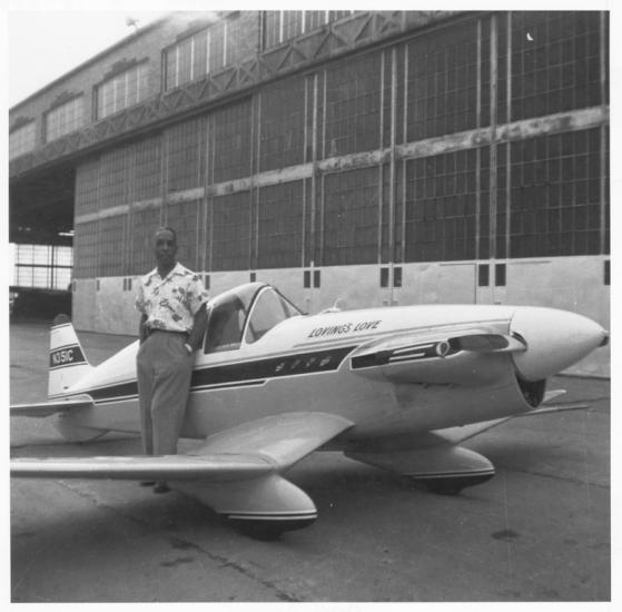 A standing person poses next to a small monoplane in front of an airplane hangar. 