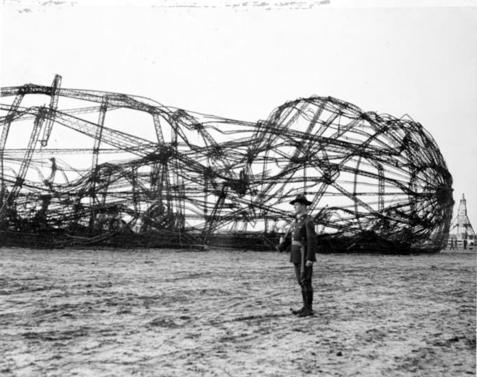 A person in uniform stands next to the wreckage of an airship.