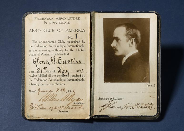 A photo of a small booklet that has been opened up to show a pilot's license. On the left page is the person's name and certification and on the right is a photo of the person's profile accompanied by their signature.