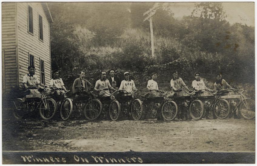 Eleven people pose with motorcycles in front of a building and bushes in this sepia-toned photo.