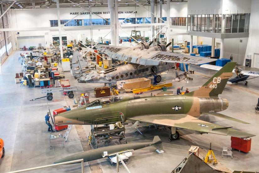 An airport hanger with two planes on the ground. They are surrounded by equipment to work on them.