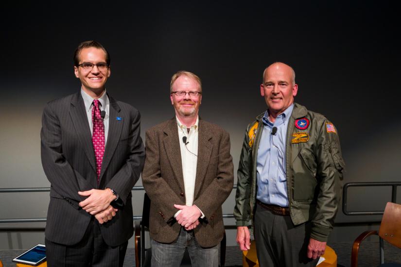 Three men smile at a camera, one wears a flight jacket. 