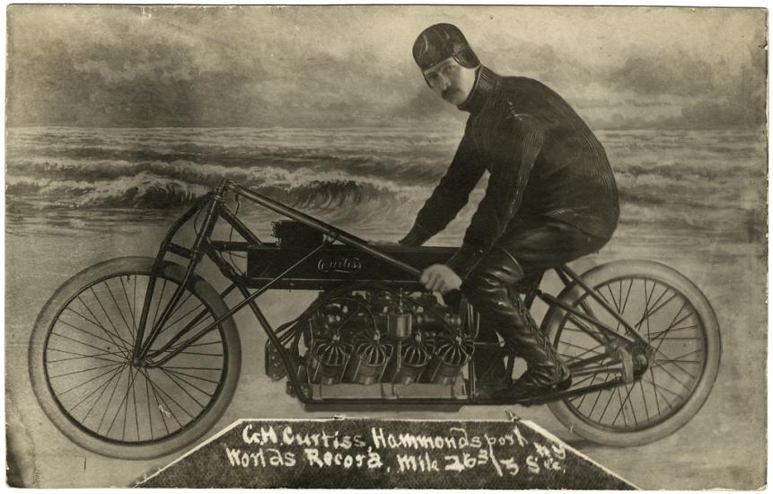 A man sits posed on a motorcycle, looking at the camera. He is in front of what is presumably a fake background meant to look like the beach.