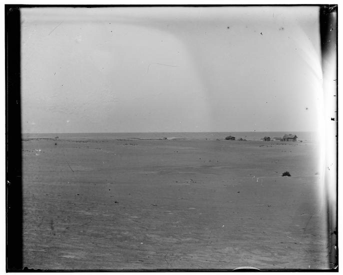 A photo in which a few small buildings can be seen in the distance. In the foreground is a flat plain. Just beyond the buildings the ocean can be seen.