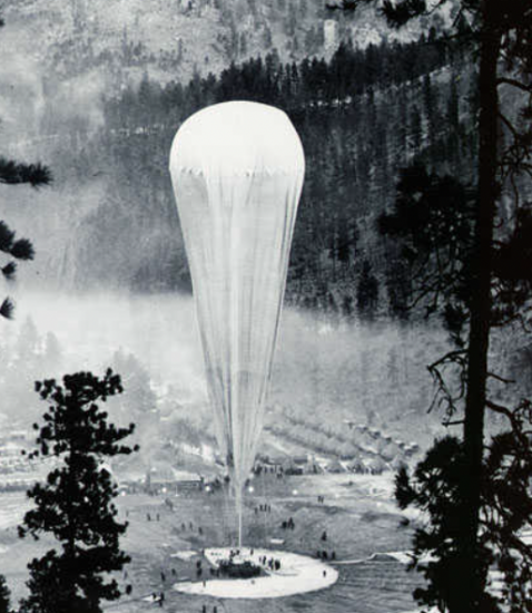 A black and white photograph of a balloon inflating above the trees. 