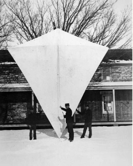 A giant four sided kite. Several men stand in front of the kite, and it is more than twice as tall as they are. 