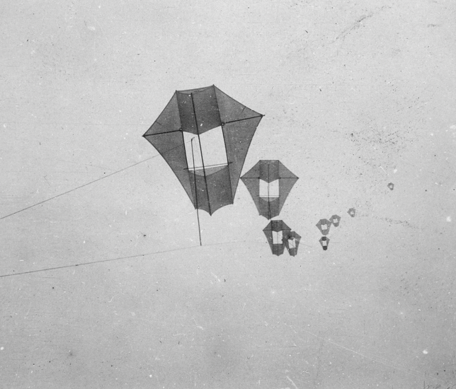 A series of kites fly, they are a polygon shape with a large rectangular opening in the middle. 