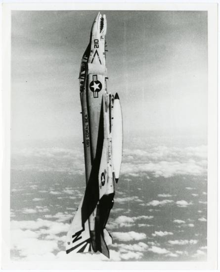 A fighter-style jet in a completely vertical position in the air. The nose of the aircraft points toward the top of the photograph and the tail toward the bottom.