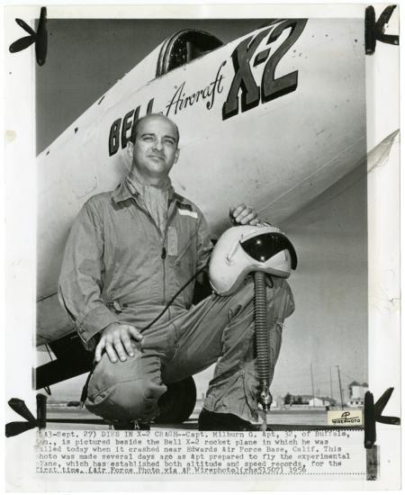 A man kneels in a flight suit in front of the nose of an aircraft which has Bell Aircraft X-2 written on the side. He balances a helmet on his knee.