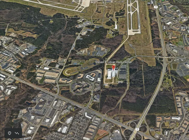 An overhead view of an aircraft hangar surrounded by a parking lot, roads, and many many trees. 