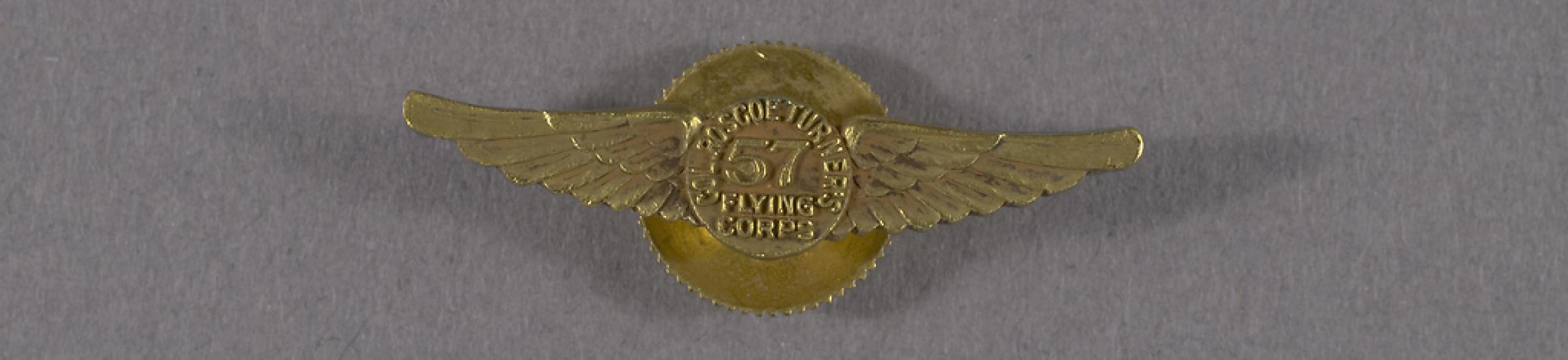 A gold pin in the shape of wings with the number 57 at the center. Around the 57 it reads Col. Roscoe Turner's Flying Corps.