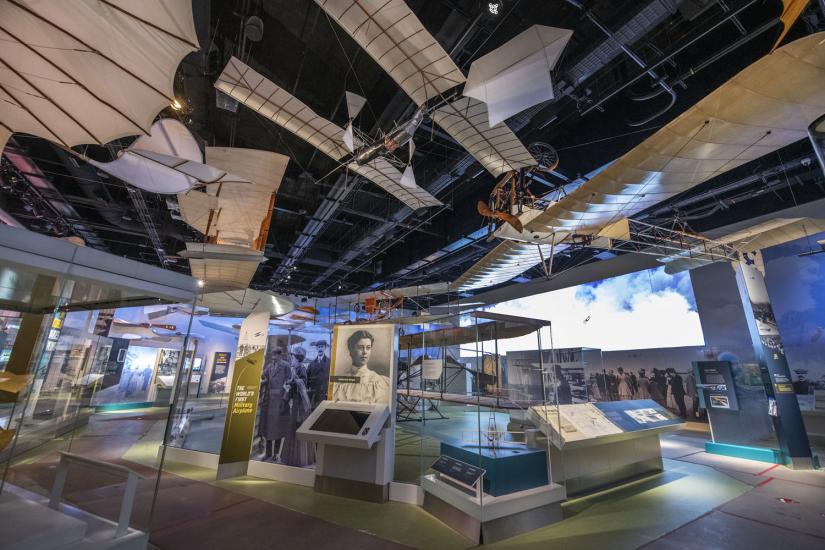 Several beige biplanes hang from the ceiling over exhibitry. 