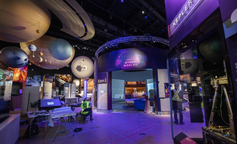 The view of a gallery from the doorway. The walls are purple. Orbs representing the planets hang above. 