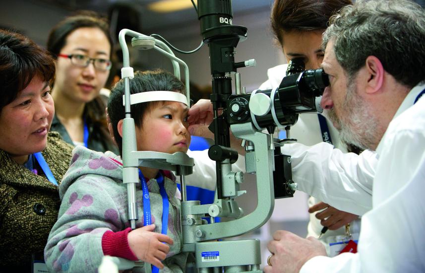 A small child looks into optometry equipment while a doctor takes measurements.