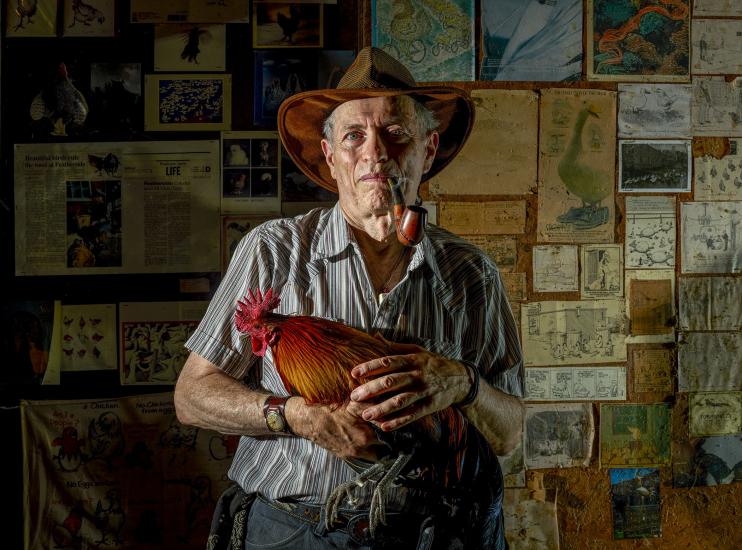 A man faces the camera holding a chicken. Behind him are various maps and newspapers hanging on the wall.