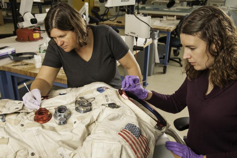 Two women next to a spacesuit working to conserve it with tools.