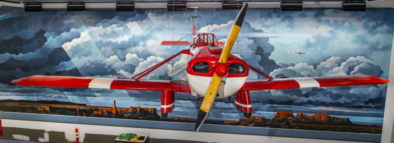 A red and white airplane hang in front of a mural of a Western landscape.