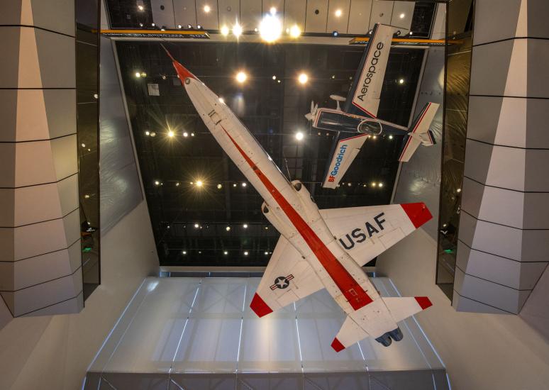 An orange and white plane with USAF written on the bottom of the wing seen from below. A smaller blue and white airplane hangs above it.