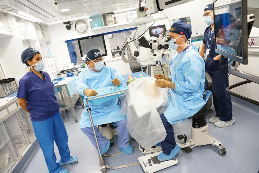 Four medical professionals stand around an operating table. One of them sits next to the table looking into a machine that is focuses on the patient.