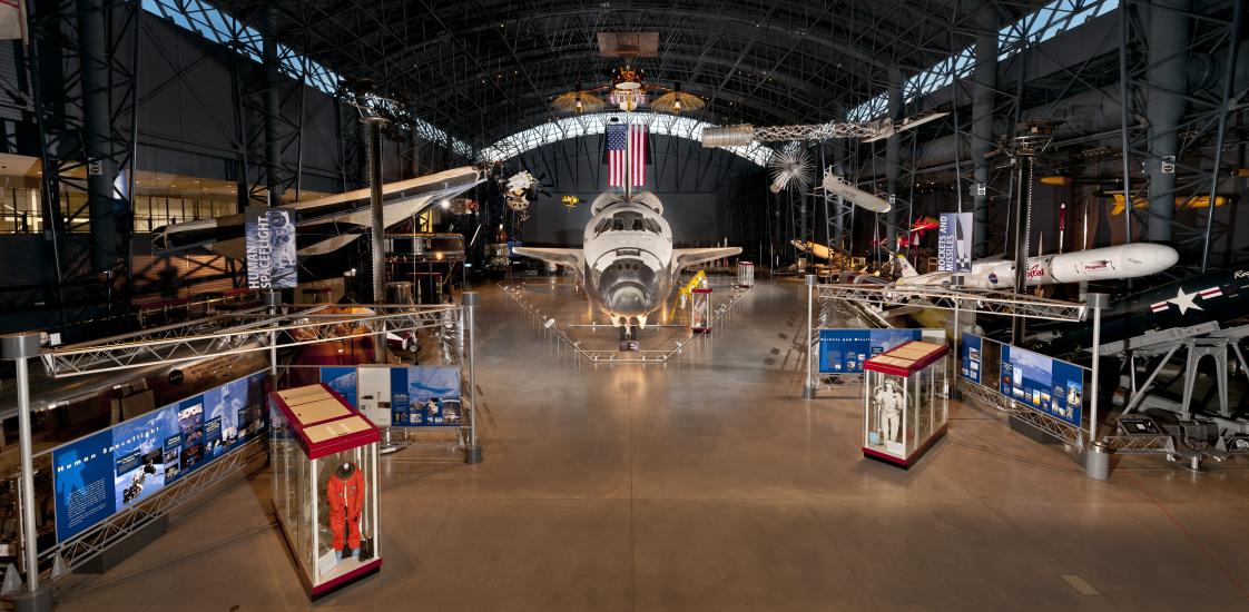 At the center of the photograph is the nose of the Space Shuttle, with the Space Shuttle expanding behind it. Hanging overhead is a satellite, a spacesuit, and a missile. Surrounding on the ground are cases with space stuff. 