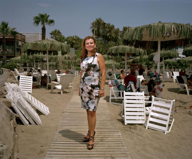 A women faces the camera, she stands on a boardwalk on the sand on the beach.