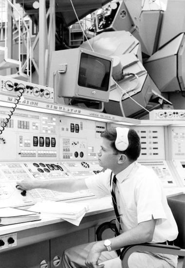 A man with a headset sits at a set of controls and computers. He appears to be flipping the position of a knob.