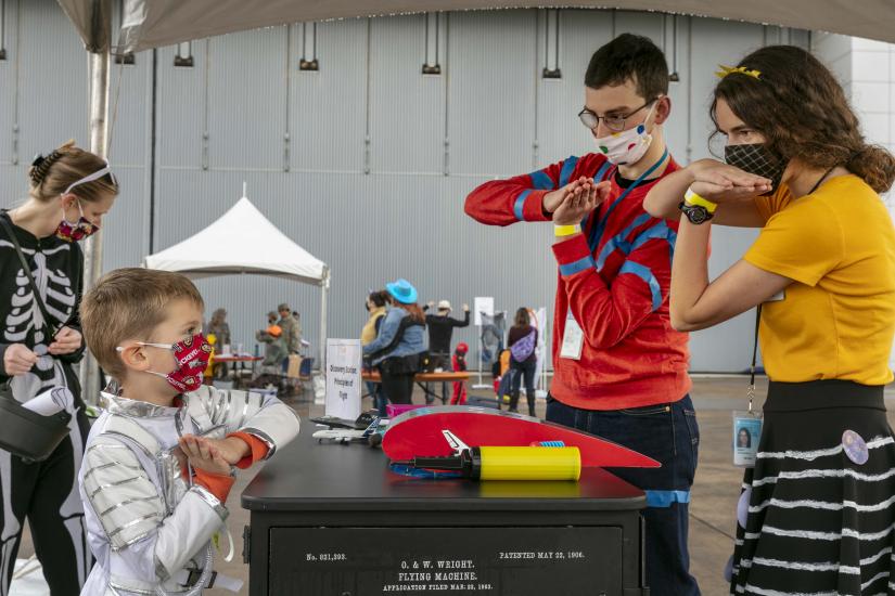 Two young adults wearing costumes demonstrate the wing of an aircraft with their hands. A young child in front of them mimics their hand motions.