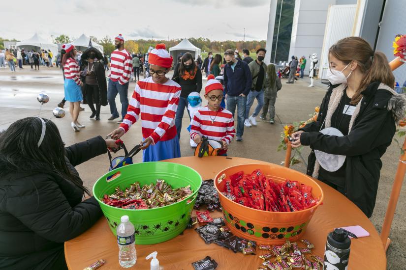 Two kids dressed as Where's Waldo trick or treat outside the Udvar-Hazy Center. There are two large tubs of candy on the tables.