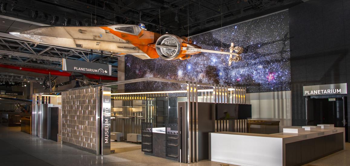 X-wing, a weathered cream and orange space vehicle hangs over an unstocked store and ticket area.