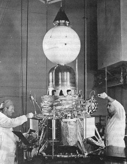 Two people stand next to a conical shaped spacecraft in a laboratory with a globe shaped feature at the top.