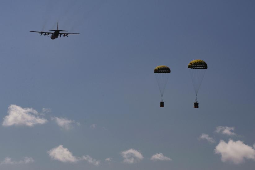 Two crates drift down by parachute with a plane in the background. 